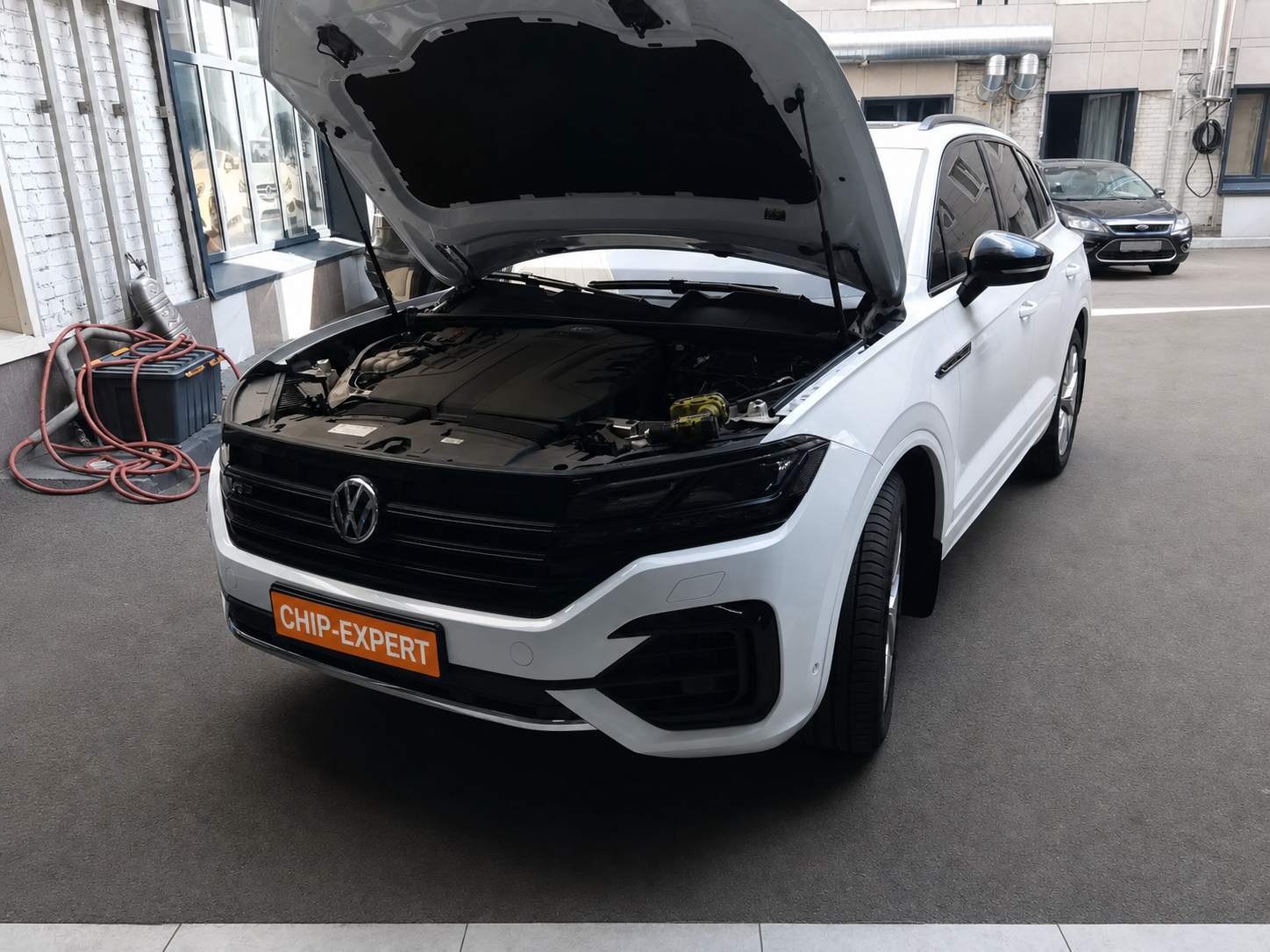 Белый Volkswagen Touareg 3.0TDI 2019 с открытым капотом в рабочей зоне автосервиса, вид спереди слева, в мастерской Чип-Эксперт — chip-expert.pro.